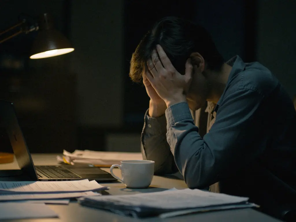 Uitgeputte persoon met hoofd in handen aan een rommelig bureau, verlicht door een enkele lamp in donkere kantooromgeving.